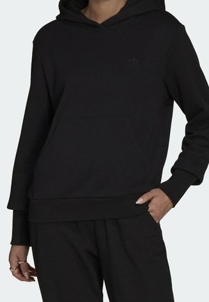 Osoba nosząca czarną bluzę z kapturem Adidas oraz pasujące czarne spodnie, z rękami w kieszeniach i widocznymi pierścieniami na palcach.