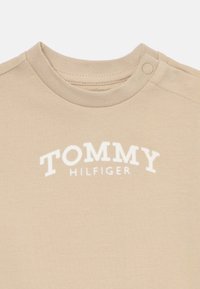 Μπεζ βαμβακερό μπλουζάκι με στρογγυλή λαιμόκοψη, που διαθέτει λευκό λογότυπο "TOMMY HILFIGER" σε όλο το στήθος και λεπτομέρεια με κουμπιά στο ώμο.