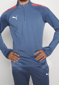 Mies sinisessä pitkähihaisessa urheilu paidassa ja housuissa, joissa on Puma-logot, kädet lanteilla, punaiset raidat hartiapaneleissa.