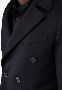 Svart ullblandad kappa med dubbelknäppt design, skarpa slag och två knappar. Har en sidficka och en mjuk textur.