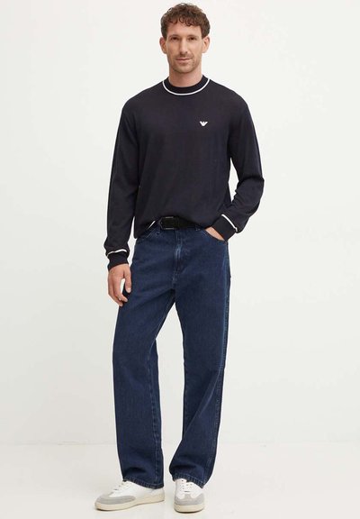 Camicia a maniche lunghe blu navy con bordino bianco al collo, abbinata a jeans in denim larghi e sneaker bianche. Presenta il logo sul petto sinistro della camicia.