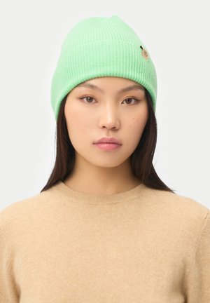 Young woman with straight dark hair wears a light green ribbed beanie and a beige crewneck sweater, facing forward against a plain background.