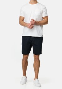 Camiseta de algodón blanca con un bolsillo en el pecho y un botón marrón, combinada con pantalones cortos azul marino. El modelo lleva zapatillas blancas. Look simple y casual.
