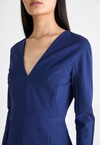 Tamsiai mėlyna suknelė ilgomis rankovėmis ir giliu V formos iškirpimu, pasižyminti elegantišku dizainu ir subtiliais siuvinėjimo akcentais ant liemenės.