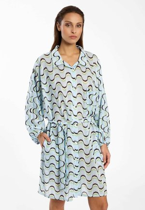 Vrouw met een knielange jurk met lange mouwen, dichtgeknoopt, met een blauwe golvende print en een ceintuur, staand met één hand in de zak.