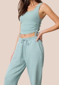 Femme portant un débardeur côtelé bleu clair avec des liens sur les côtés et un pantalon de survêtement à cordon assorti, debout avec la main sur la hanche sur un fond uni.
