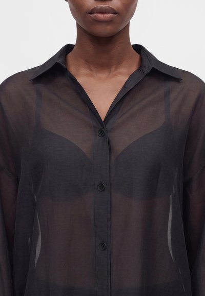 Personne portant une chemise noire transparente boutonnée, laissant apparaître un soutien-gorge noir en dessous, montrée du cou jusqu'au milieu du torse sur un fond uni.