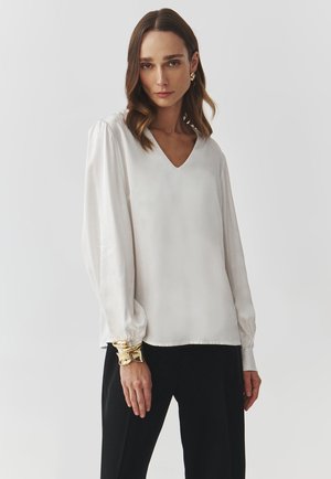 Blusa di seta bianca con scollatura a V, maniche lunghe a sbuffo e polsini arricciati, abbinata a pantaloni neri e un bracciale dorato come accentuazione.
