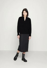 Kvinne i svart langermet topp, svart midi-skjørt og svarte lær ankelstøvler, som står mot en enkel lys bakgrunn.