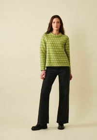 Kvinna står iklädd en grön mönstrad tröja, svarta vida byxor och svarta skor mot en enfärgad beige bakgrund.