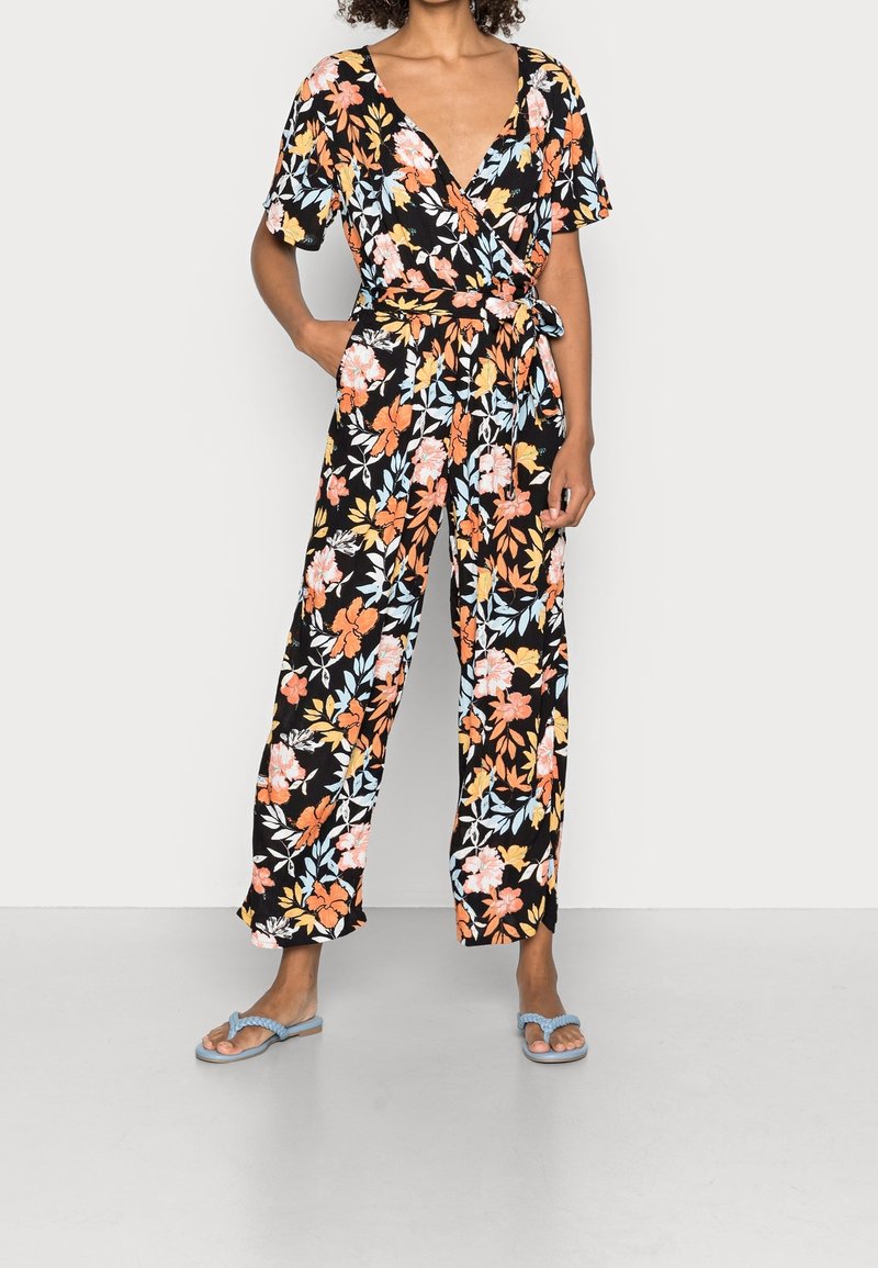 Combinaison fleurie à base noire, ornée de fleurs orange, rose et bleu clair. Col en V, manches courtes, taille nouée et coupe à jambes larges.
