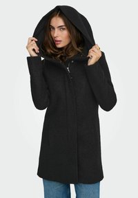 Manteau noir à capuche en tissu texturé et épais, avec une fermeture éclair à l'avant et des poches latérales, partiellement zippé, porté par un mannequin.