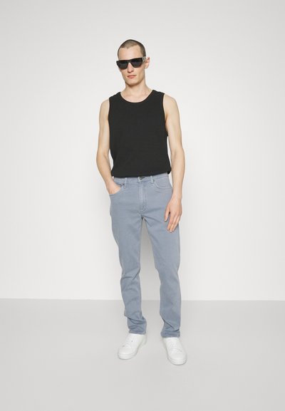 Black sleeveless shirt, light blue jeans, and white sneakers. Model wearing dark sunglasses, with a minimalist background.