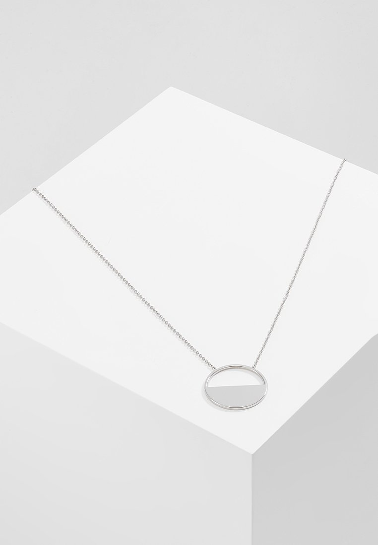 Collier en chaîne argentée avec un pendentif circulaire présentant un motif horizontal à moitié rempli, exposé sur une surface géométrique blanche.