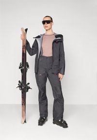 Black waterproof ski jacket and pants, textured light grey sweater, black ski boots, holding a pair of skis, oversized black sunglasses.
