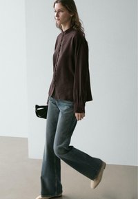 Braunes Hemd mit Knopfleiste und hohem Kragen, kombiniert mit ausgestellten blauen Jeans und beigefarbenen Schuhen. Verfügt über eine entspannte Passform und sichtbare Nähte als Akzente.