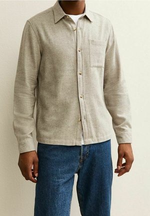 Mann trägt ein beige-farbenes Hemd mit Knöpfen und Brusttasche über einem weißen Unterhemd, kombiniert mit blauen Jeans vor einer schlichten Wand.