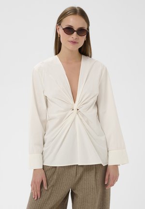 Žena nosiace okrúhle tmavé slnečné okuliare, krémovú blúzku so zvrúteným uzlom a hlbokým výstrihom do V a hnedé široké nohavice s pruhmi.