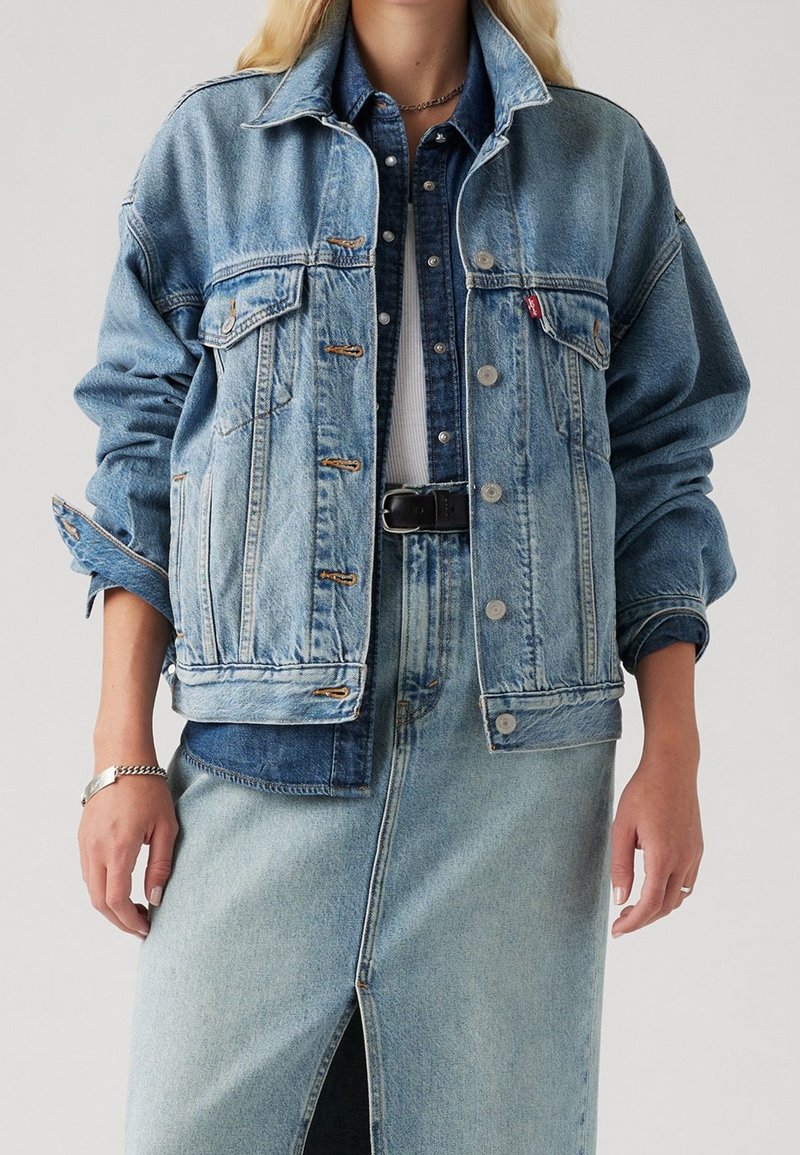 Veste en denim bleu clair avec une coupe décontractée, grandes poches avant, fermetures à boutons et accents bleus contrastants, portée sur une jupe en denim.