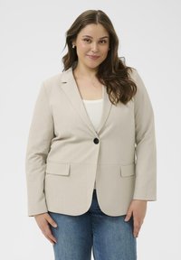 Beige räätälöity bleiseri, jossa on yksittäinen nappisulku, viistot läpät ja kaksi etutaskua, yhdistettynä valkoiseen yläosaan ja sinisiin farkkuihin.