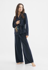 Conjunto de pijama de terciopelo azul marino que presenta un diseño de top cruzado con bordado de dragón y ribetes en rosa contrastante, complementado con pantalones de pierna ancha.