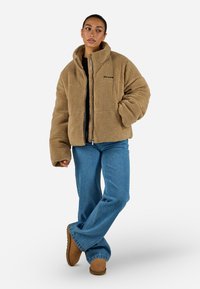 Giacca in pile marrone con colletto alto, chiusura a zip e vestibilità over. Indossata sopra una maglietta nera, abbinata con jeans blu e stivali color nocciola.