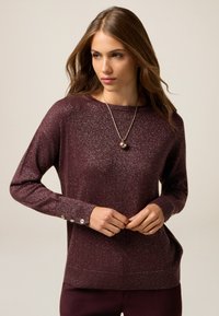 Maglione bordeaux con una morbida texture scintillante. Presenta maniche lunghe e dettagli in bottone sui polsi. Collo a giro, vestibilità morbida.