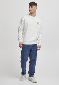 Hvid sweatshirt med rund halsudskæring og broderet logo på brystet, parret med blå denim jeans og hvide sneakers.