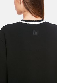 Schwarzes Sweatshirt mit einem gewellten, weiß gestreiften Kragen, auf der Rückseite ist ein dezentes Logo. Weicher Stoff, lockere Passform, minimalistisches Design.