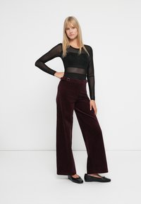 Maglietta nera trasparente a maniche lunghe abbinata a pantaloni a gamba larga in corda bordeaux con passanti per cintura. Il modello indossa scarpe nere basse.