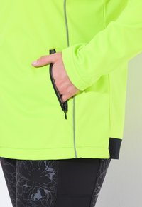 Fluorescentti vihreä takki pitkine hihoineen ja vetoketjullisella taskulla; sisältää kontrastiväriset mustat yksityiskohdat ja tekstuurilla varustetut mustat legginsit, joissa on kukkakuosi.