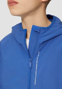 Blaue Zip-Jacke mit Kapuze, glatter Textur und einem weißen Drucktextdetail auf der Vorderseite. Reißverschluss ist sichtbar und funktional.