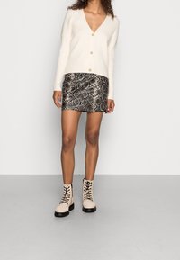 Cremfarbener V-Ausschnitt-Cardigan mit geripptem Muster, kombiniert mit einem Minirock im Schwarz-Silber-Schlangenprint. Beigefarbene Schnür-Stiefeletten mit schwarzer Sohle.