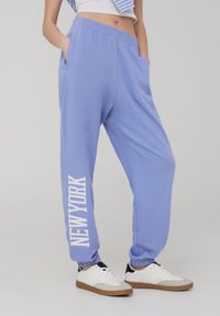Pantaloni della tuta azzurro chiaro con vita elastica e caviglie a coste; presenta la scritta "NEW YORK" stampata in bianco bold sulla gamba sinistra.
