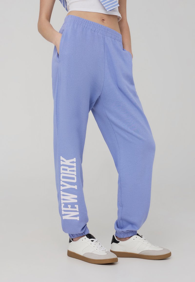Pantaloni della tuta azzurro chiaro con vita elastica e caviglie a coste; presenta la scritta "NEW YORK" stampata in bianco bold sulla gamba sinistra.