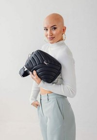 Černá prošívaná crossbody taška se stříbrnými prvky, na které se nachází logový akcent. Model má na sobě bílý svetr a světle modré kalhoty.