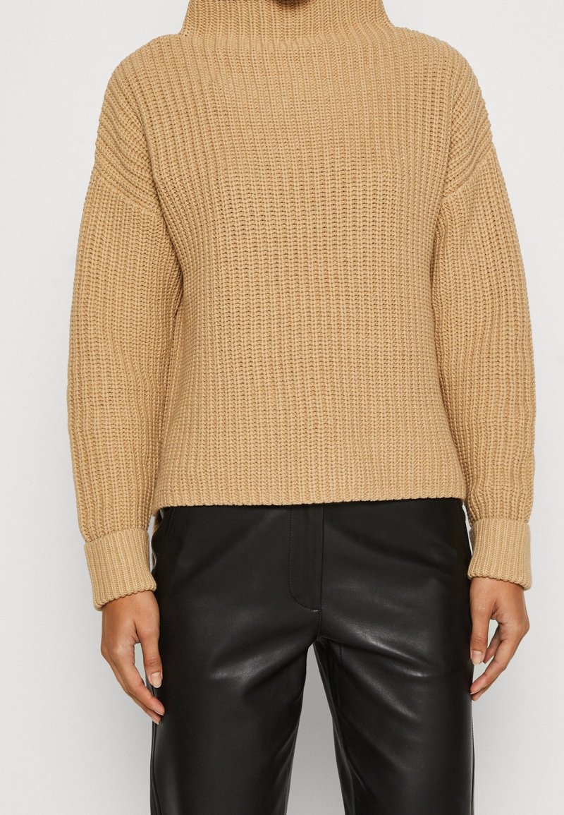 Personne portant un pull à col roulé en maille épaisse beige et un pantalon en cuir noir sur un fond uni.