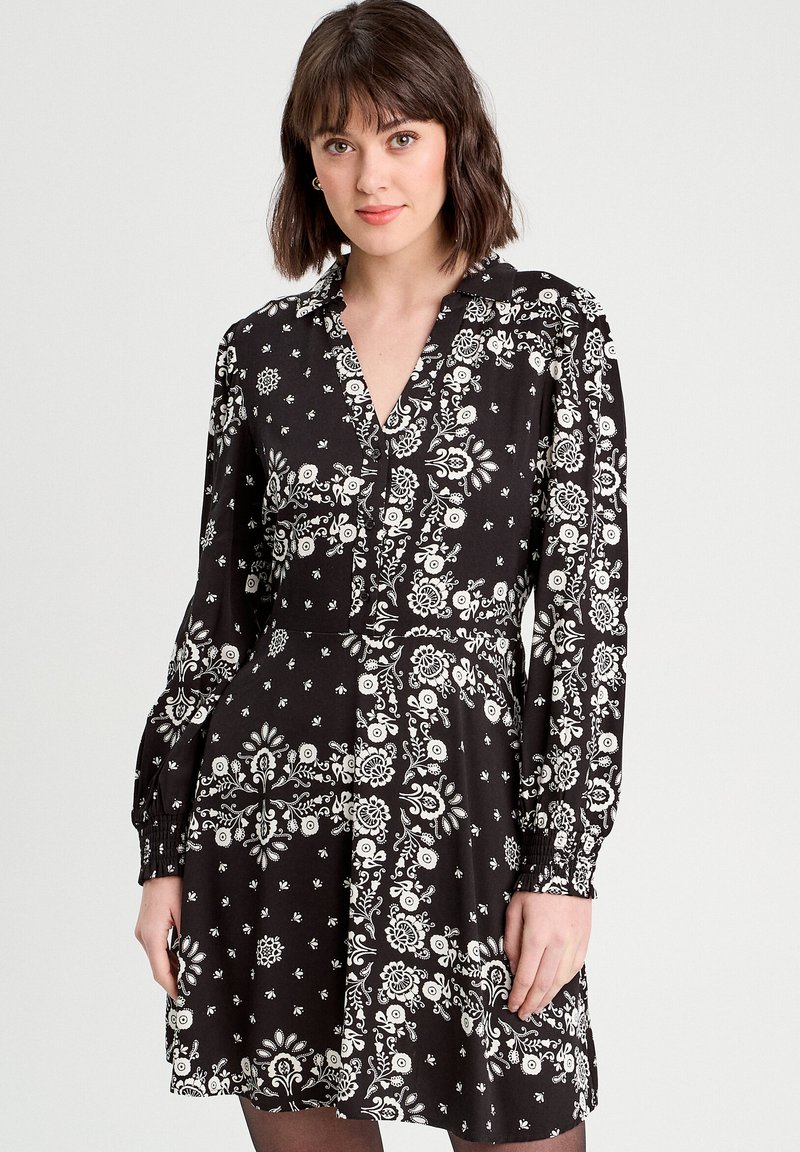 Robe noire avec des motifs floraux blancs, fermeture à boutons sur le devant et longues manches bouffantes. Présente un corsage ajusté et une jupe évasée. Tissu léger.