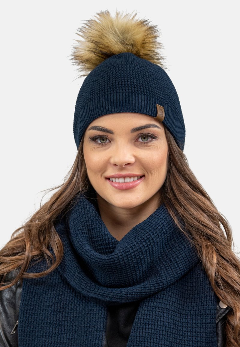 Căciulă tricotată navy cu un pompon din blană sintetică în vârf. Șalul asociat are un design texturat, cu dungi, tot în nuanța navy.