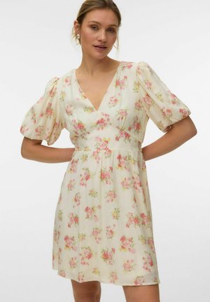 Mujer que lleva un vestido corto crema con estampado floral rosa y verde, mangas cortas abullonadas y escote en V, posando con las manos en las caderas.