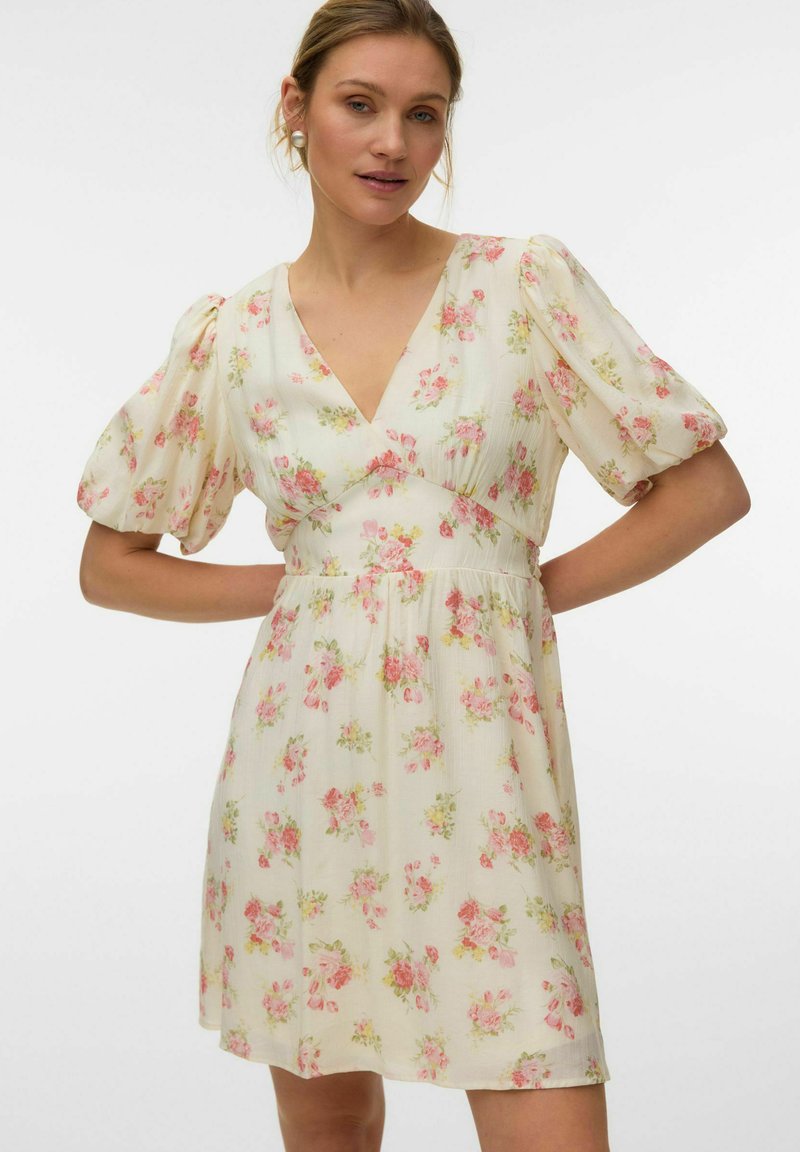 Femme portant une mini-robe crème à imprimé floral rose et vert, avec des manches courtes bouffantes et un décolleté en V, posant avec les mains sur les hanches.