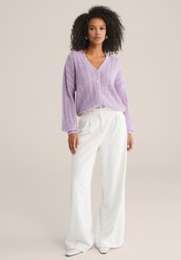 Violeta adīta kardigāna ar V-veida kaklu, priekšpuses pogu aizdari un brīviem gariem piedurknēm, kopā ar baltām platām biksēm un augstpapēžu kurpēm.