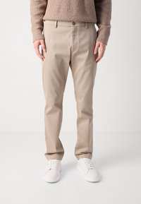 Beige Chino-Hosen mit geradem Bein, aus Baumwolle. Verfügt über einen Knopfverschluss, Seitentaschen und wird mit weißen Sneakers getragen.