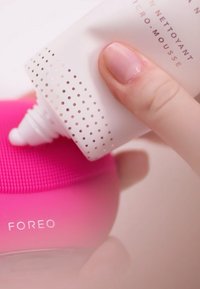 Rosa silikon ansiktsrenseapparat med strukturert overflate; en hvit tube med rosa prikket design blir klemt over den.