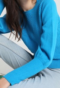 Femme portant un pull en maille texturée bleu vif et un jean en denim clair, assise avec un bras reposant sur son genou.