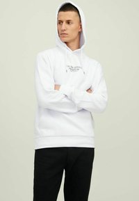 Weiße Kapuzenjacke aus Baumwollmischung mit einem Logo-Print auf der Vorderseite, verstellbaren Kordeln und einer Kängurutasche. Kombiniert mit schwarzen Hosen.
