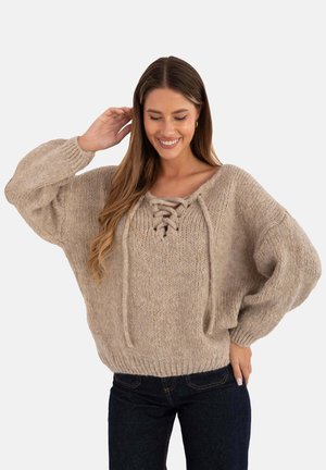 Beżowy sweter dzianinowy o luźnym kroju, szerokich rękawach, dekolcie sznurowanym oraz fakturowanej tkaninie. Noszony z ciemnymi dżinsami.