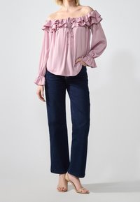 Blusa leggera rosa pallido, con scollatura off-the-shoulder e dettagli arricciati, maniche lunghe e scollatura con stringhe, abbinata a jeans a gamba larga scuri e tacchi beige.
