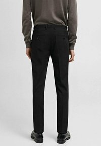 Schwarze Hose mit glatter Textur, schmaler Passform und Gürtelschlaufen. Verfügt über zwei Gesäßtaschen mit Knöpfen. Kombiniert mit dunklen Schuhen.