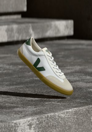 Zapatilla blanca con detalles verdes, presenta una puntera texturizada de ante y una suela de goma. Diseño con cordones y un prominente logotipo en forma de "V".